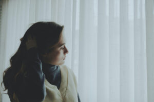 A woman looks anxious next to a window curtain.